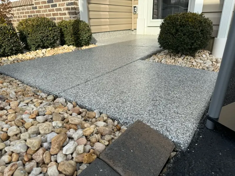 Concrete walkway leads to a home's entrance, bordered by small stones and neatly trimmed bushes, with contrasting siding and bricks.