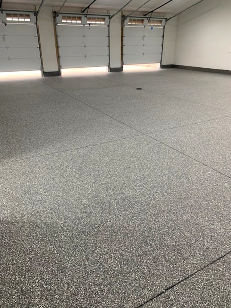 A spacious, empty garage with a clean epoxy floor and closed white panel doors. Overhead lighting softly illuminates the well-organized space.