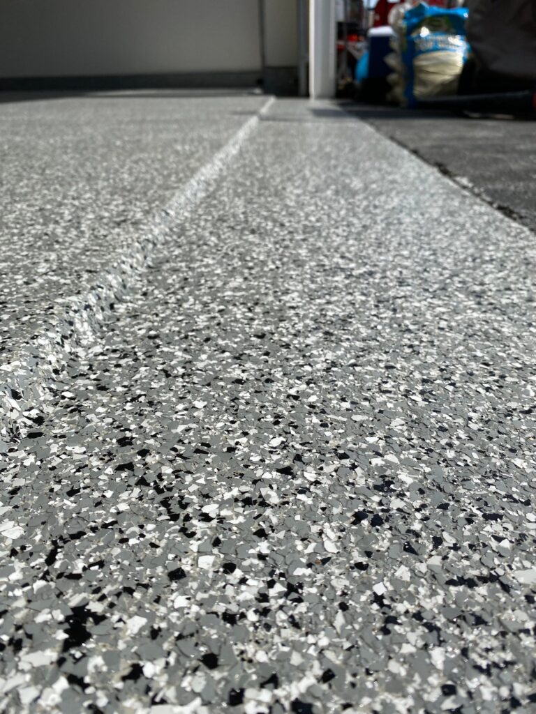 Close-up view of a textured, speckled garage floor with a small portion of an open garage entrance and storage items visible in the background.