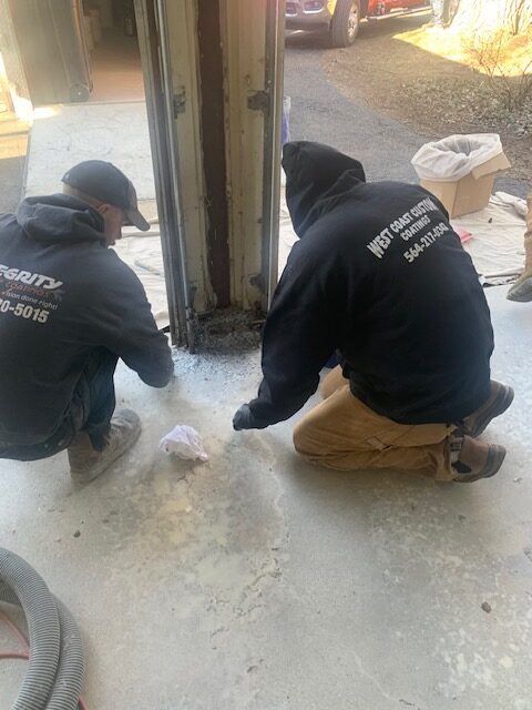 Two persons wearing dark hoodies work on a concrete floor near a garage door. A cardboard box and vehicle are visible nearby.
