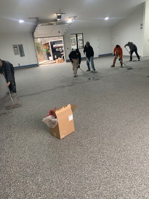 Five people are working on resurfacing a large indoor floor. A cardboard box is in the foreground. The garage door is open.