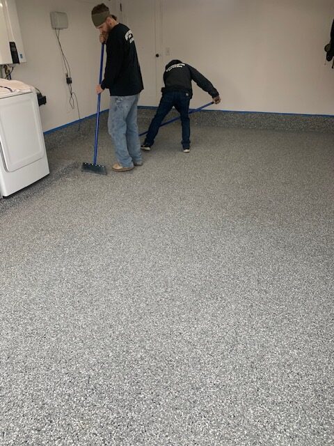 Two people installing a speckled epoxy floor in a room with a washing machine. One person uses a brush, the other smooths the surface.